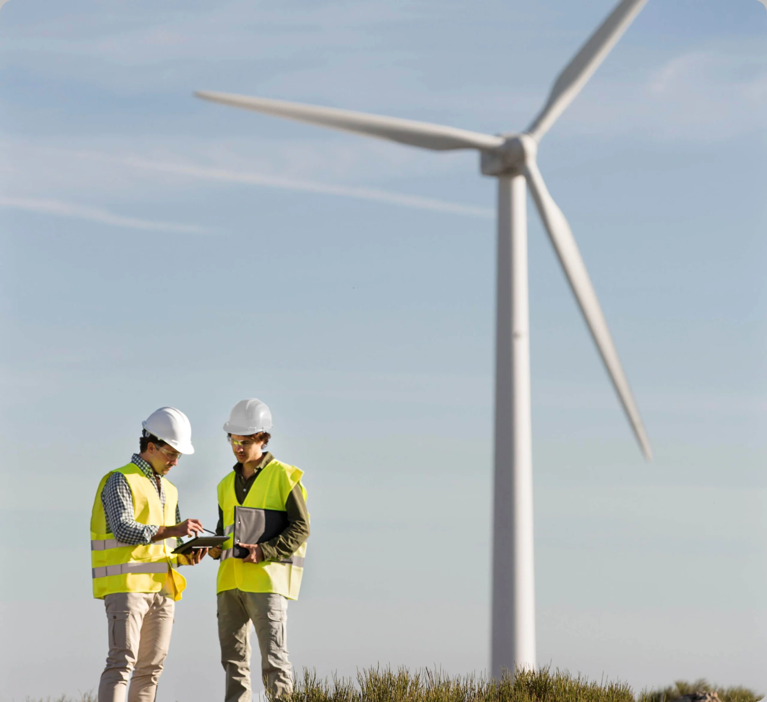 Deux travailleurs portant des gilets de sécurité et des casques se tiennent devant une grande éolienne. L’un tient une tablette et semble discuter de quelque chose tout en examinant l’appareil. Le ciel est dégagé et le décor parais être un parc éolien.