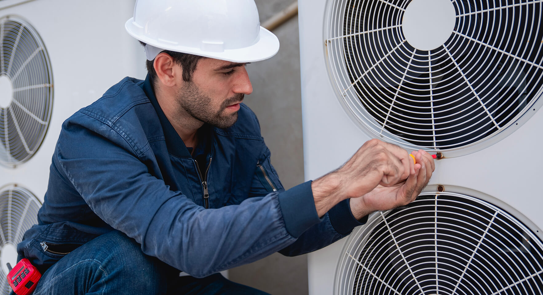 Un homme portant un casque blanc et une veste bleue travaille sur une grande unité de climatisation. Il utilise un tournevis pour effectuer des réglages sur l'unité, avec d'autres outils visibles dans sa ceinture à outils. L'arrière-plan est neutre et flou.